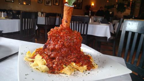 A plate of pasta topped with a large mound of meat sauce and a bone, garnished with herbs and cheese.