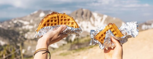 Two hands hold waffles wrapped in foil against a scenic mountain backdrop.