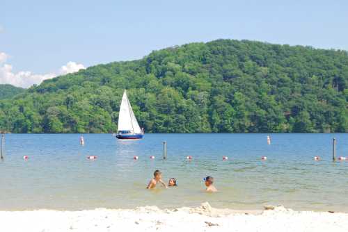 Three children play in shallow water at a beach, with a sailboat in the background and green hills beyond.