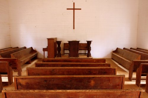 Interior of a simple church with wooden pews, a cross on the wall, and a small altar at the front.