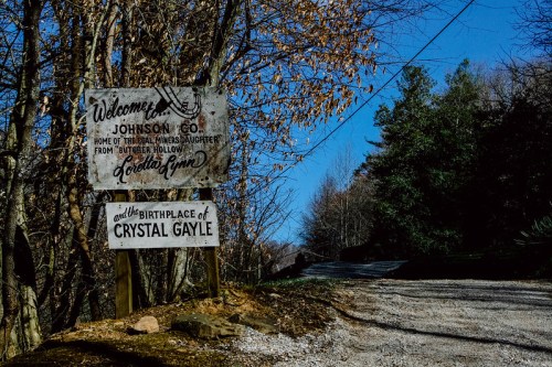 A weathered sign welcomes visitors to Johnson County, birthplace of Crystal Gayle, surrounded by trees and a gravel road.