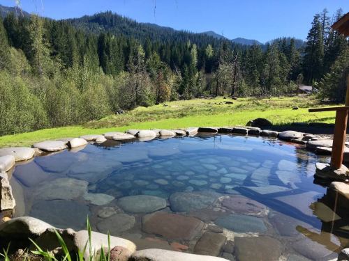A serene hot spring surrounded by lush greenery and mountains under a clear blue sky.