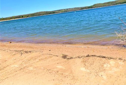 A serene lakeshore with sandy beach, gentle waves, and green hills in the background under a clear blue sky.