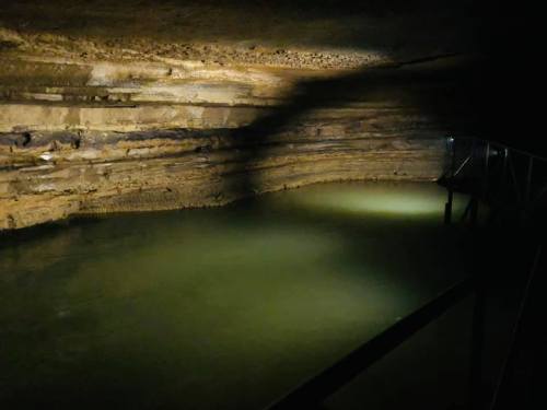 A dimly lit cave with layered rock formations and a still body of water, featuring a metal walkway.