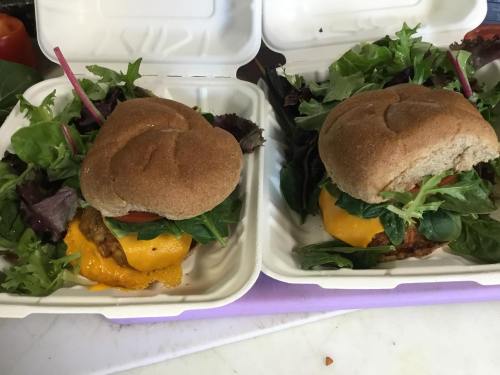 Two takeout containers with whole grain burgers topped with cheese, lettuce, and tomato, surrounded by fresh greens.