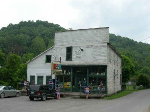 A rustic white building with a porch, people gathered outside, surrounded by green hills and trees.