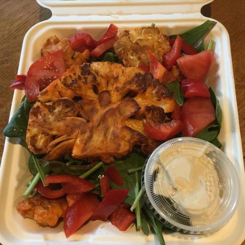 A takeout container filled with leafy greens, sliced tomatoes, and a large piece of roasted cauliflower.