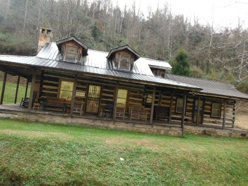 A rustic log cabin with a metal roof, featuring a porch and surrounded by trees and grassy land.