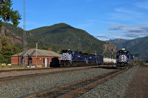 Two blue and white locomotives on tracks near a brick train station, with mountains in the background under a clear sky.