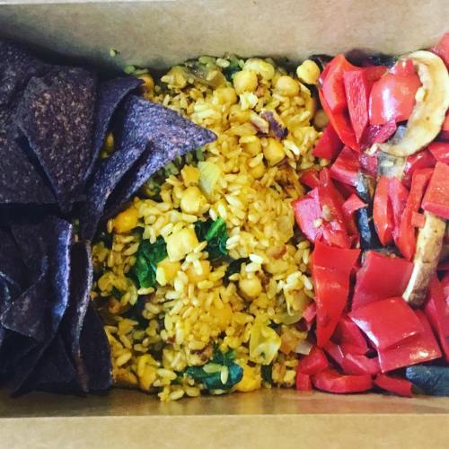 A takeout box with blue corn chips, rice with corn and greens, and mixed red and yellow bell peppers.