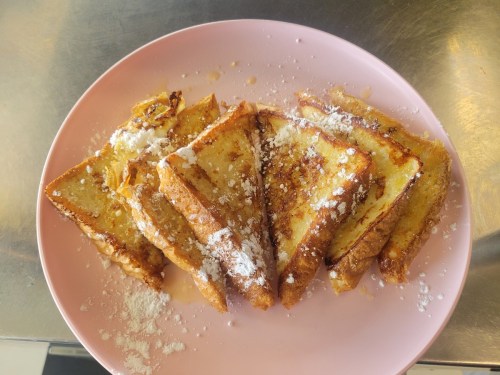 A pink plate with several slices of golden-brown French toast, dusted with powdered sugar and drizzled with syrup.