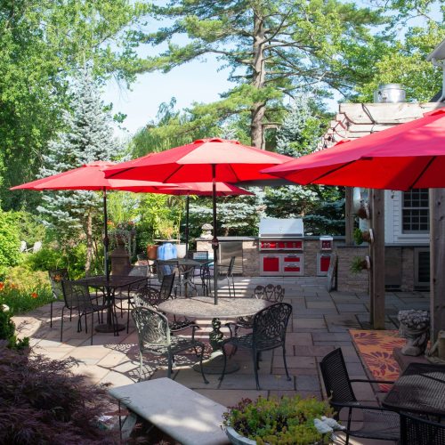 A sunny outdoor patio with red umbrellas, tables, and a grill surrounded by lush greenery and trees.