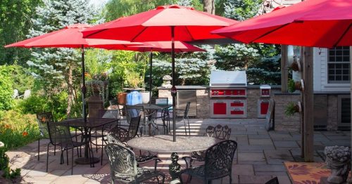 A patio with red umbrellas over metal tables, surrounded by greenery and featuring a grill in the background.