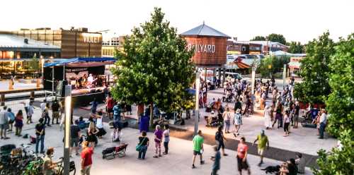 A lively outdoor event with a crowd enjoying a performance, surrounded by trees and buildings at dusk.