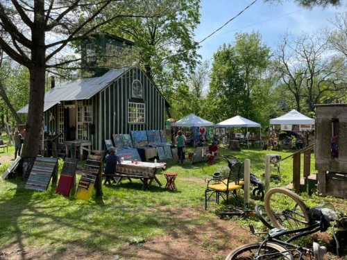 A vibrant outdoor art market with tents, paintings, and a rustic building surrounded by greenery.