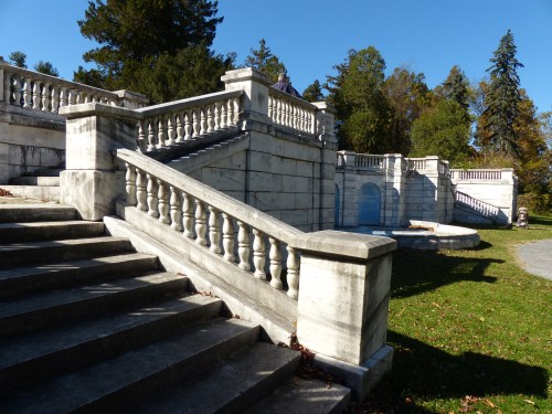 A marble staircase with ornate railings leads to a landscaped area surrounded by trees under a clear blue sky.