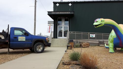 A blue truck parked outside the Great Plains Dinosaur Museum, featuring a colorful dinosaur statue by the entrance.