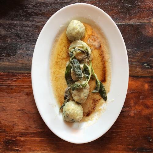 A white plate with four green dumplings in a buttery sauce, garnished with sage leaves, on a wooden table.
