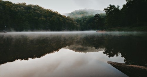 A serene lake surrounded by misty hills and lush trees, reflecting the calm water's surface.