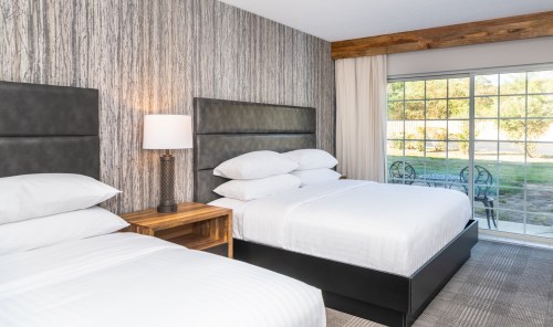 A modern hotel room with two beds, a nightstand, and a sliding glass door leading to an outdoor area.