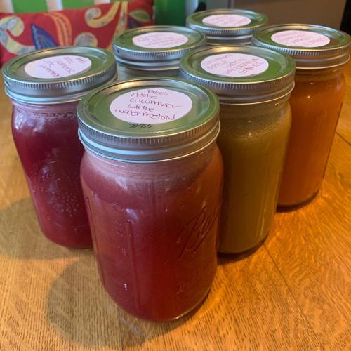 Six jars of colorful homemade beverages sit on a wooden table, labeled with flavors like watermelon and apple.