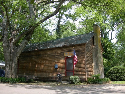 A small wooden building with a flag, surrounded by trees and benches in a park-like setting.