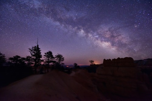 A starry night sky filled with the Milky Way over a rugged landscape with silhouetted trees and rock formations.