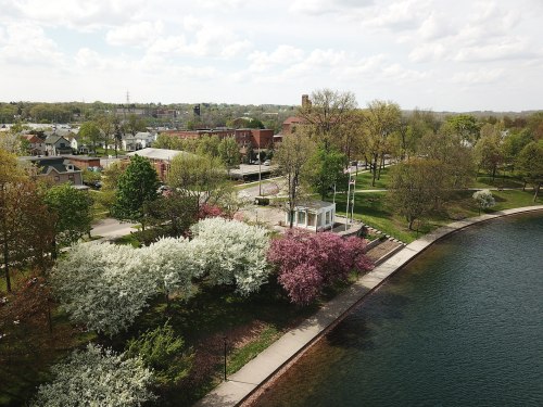 Aerial view of a park with blooming trees, a gazebo, and a calm water body, surrounded by a small town.