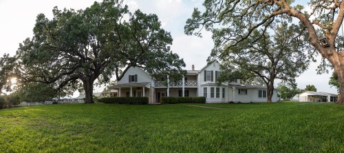 A large, two-story house surrounded by green grass and trees, with a porch and a peaceful outdoor setting.