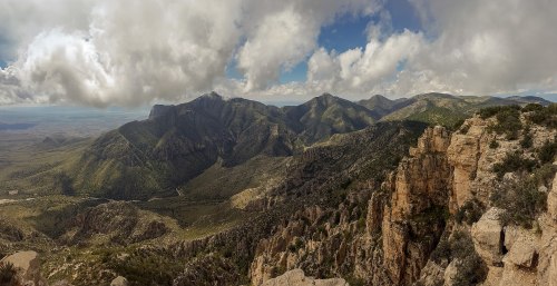 A panoramic view of rugged mountains and valleys under a partly cloudy sky, showcasing lush greenery and rocky cliffs.