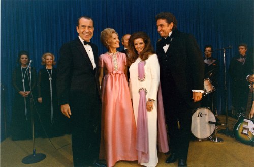 A group of four people in formal attire, smiling together on stage, with musicians and an audience in the background.