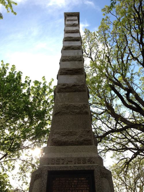 A tall, stone monument with a textured surface, surrounded by trees and a bright sky in the background.