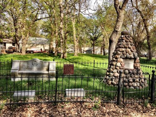 A peaceful cemetery scene with a stone monument and a bench, surrounded by trees and a black fence.