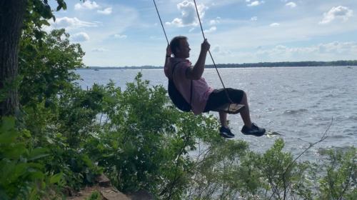 A person swings over a river, surrounded by greenery and under a blue sky with fluffy clouds.