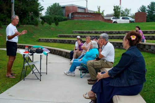A speaker addresses a small audience seated on an outdoor amphitheater, surrounded by greenery and buildings.