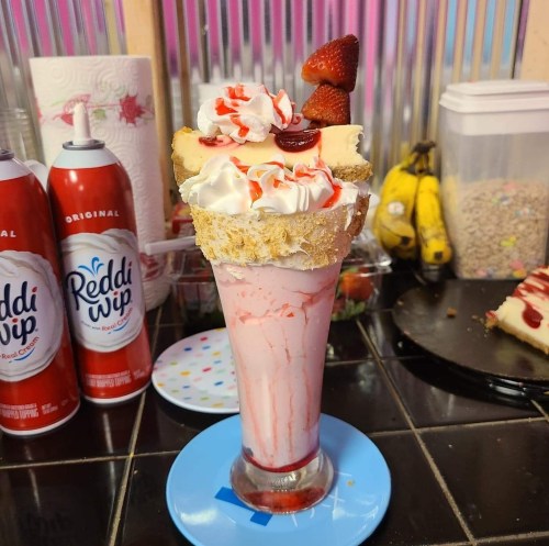 A tall dessert glass filled with pink milkshake, topped with whipped cream, strawberries, and a slice of cake.
