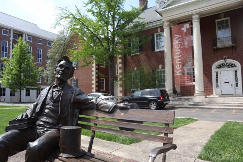 A bronze statue of Abraham Lincoln sitting on a bench in front of a brick building with a Kentucky banner.