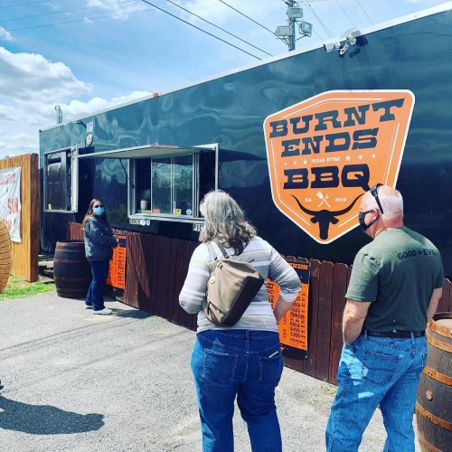 A food truck named "Burnt Ends BBQ" with customers waiting in line, set against a cloudy sky.