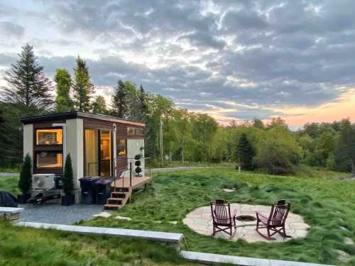 A tiny house on a grassy area with a stone fire pit and chairs, surrounded by trees under a cloudy sky at sunset.