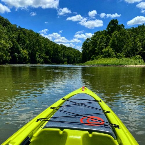 A kayak on a calm river surrounded by lush green trees and a bright blue sky with fluffy clouds.