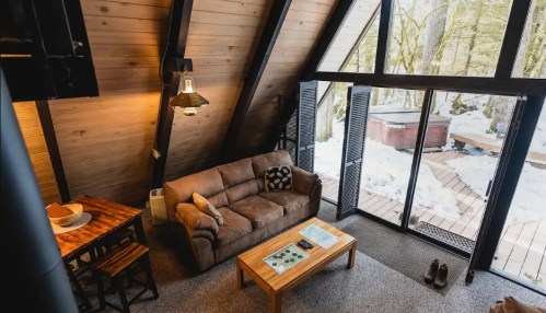 Cozy A-frame cabin interior with a sofa, wooden table, and large windows overlooking a snowy landscape.