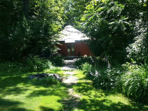 A red cabin nestled among lush greenery, with a pathway leading to it and a fire pit in the foreground.
