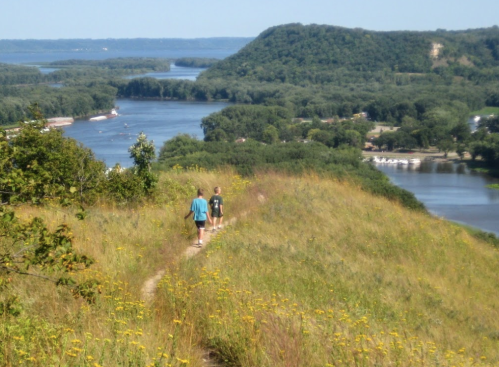 Two hikers walk along a grassy trail overlooking a river and lush green landscape on a sunny day.