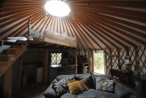 Cozy interior of a yurt with a loft, wooden beams, and a large circular skylight. Soft seating and rustic decor visible.