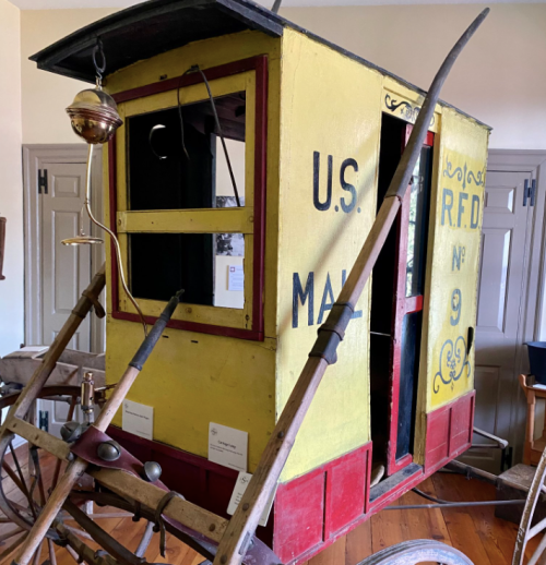 Historic yellow mail cart with red accents, labeled "U.S. Mail R.F.D. No. 9," displayed in a museum setting.