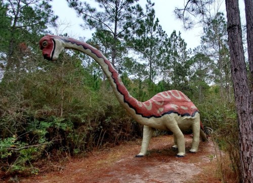A large, colorful dinosaur statue stands among trees in a wooded area, showcasing a long neck and distinctive patterns.
