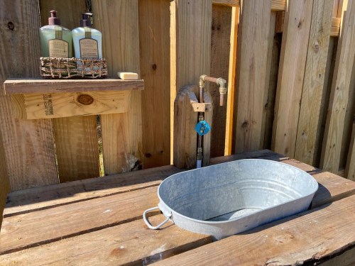 A rustic outdoor sink setup with a galvanized tub, wooden shelf, and bottles on a wooden fence background.