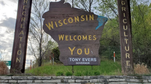 Wooden sign welcoming visitors to Wisconsin, featuring the state outline and the name of Governor Tony Evers.