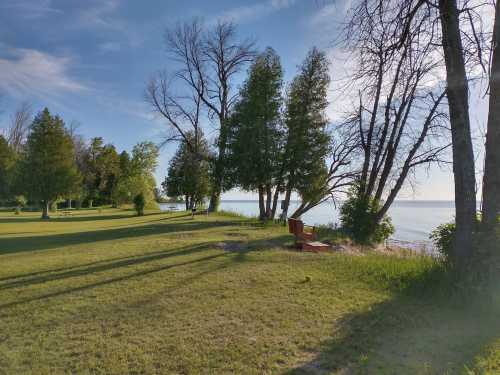 A serene lakeside scene with green grass, trees, and a red chair overlooking the water under a clear blue sky.