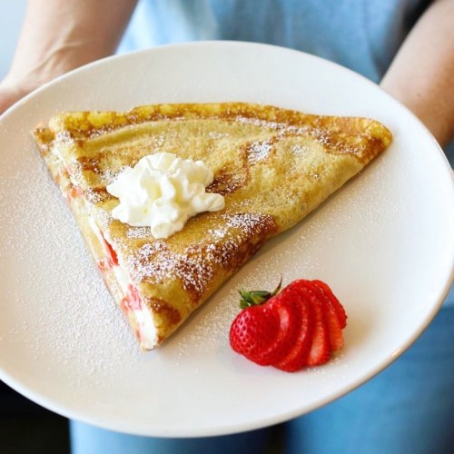 A person holds a plate with a triangular crepe topped with whipped cream and a fresh strawberry, dusted with powdered sugar.